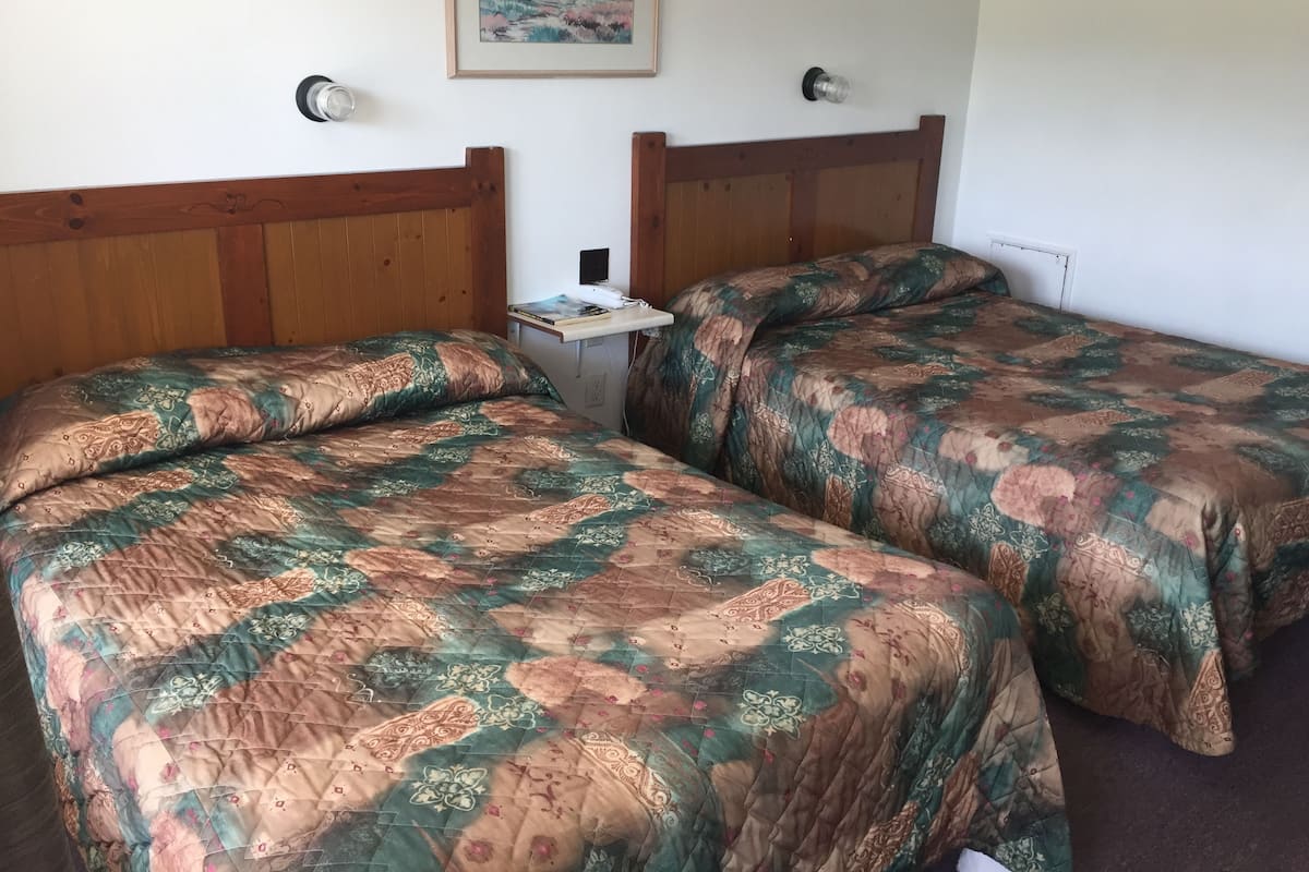 A room with two wooden beds, each with a floral-patterned bedspread. There is a small table with a lamp and a framed picture on the wall above the beds.