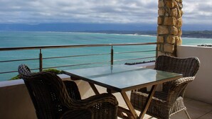 Un balcon agrémenté de fauteuils en osier et d’une table en verre, offrant une vue sur une mer turquoise et des montagnes lointaines.