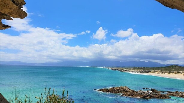 Un paysage côtier avec une eau bleue et limpide, des plages de sable et des montagnes lointaines sous un ciel partiellement nuageux.