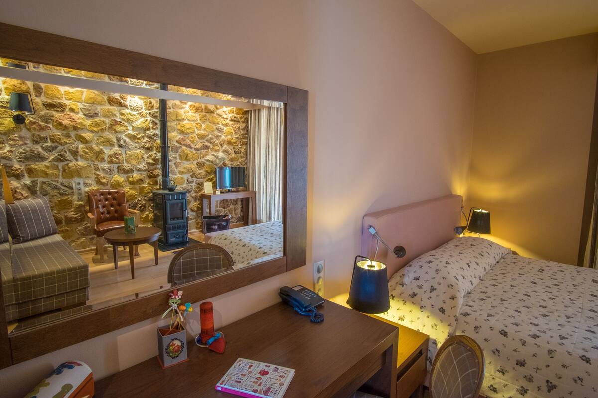 Quarto de hotel acolhedor com parede de pedra, cama com colcha floral, escrivaninha de madeira, uma cadeira, uma mesinha e um espelho refletindo o quarto.