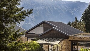 Un chalet de montagne à la façade en pierre, doté de grandes fenêtres et d’un toit en métal.