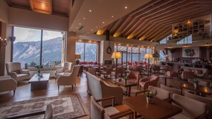 Un vaste espace de détente avec de nombreux sièges, de grandes fenêtres offrant une vue sur les montagnes et un plafond haut avec des poutres apparentes.