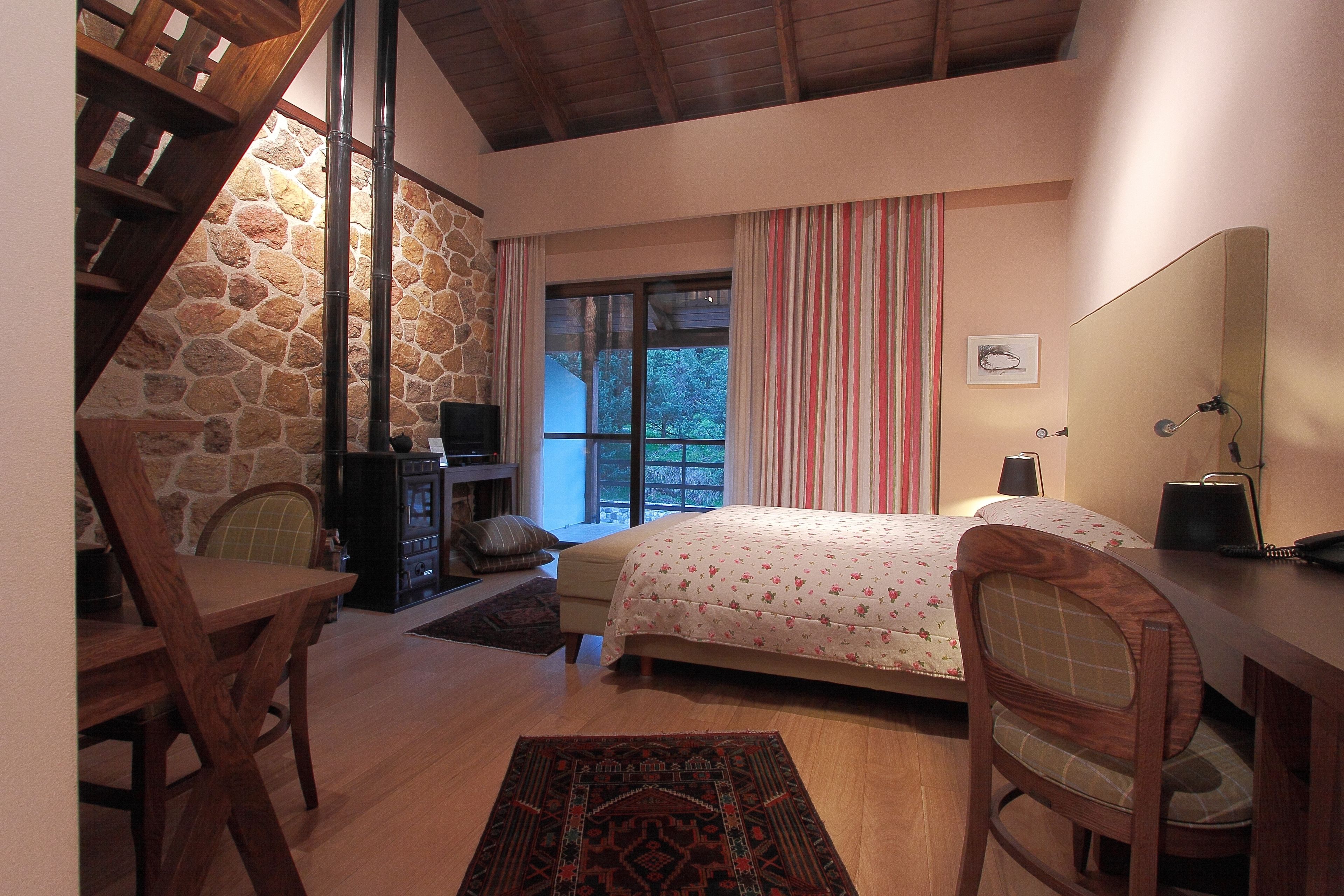 A bedroom with a stone wall, a bed with floral bedding, a desk, and a chair.