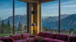 Une pièce dotée d’une grande fenêtre donnant sur les montagnes, d’un canapé violet, d’une table avec une lampe et d’un vase décoratif.