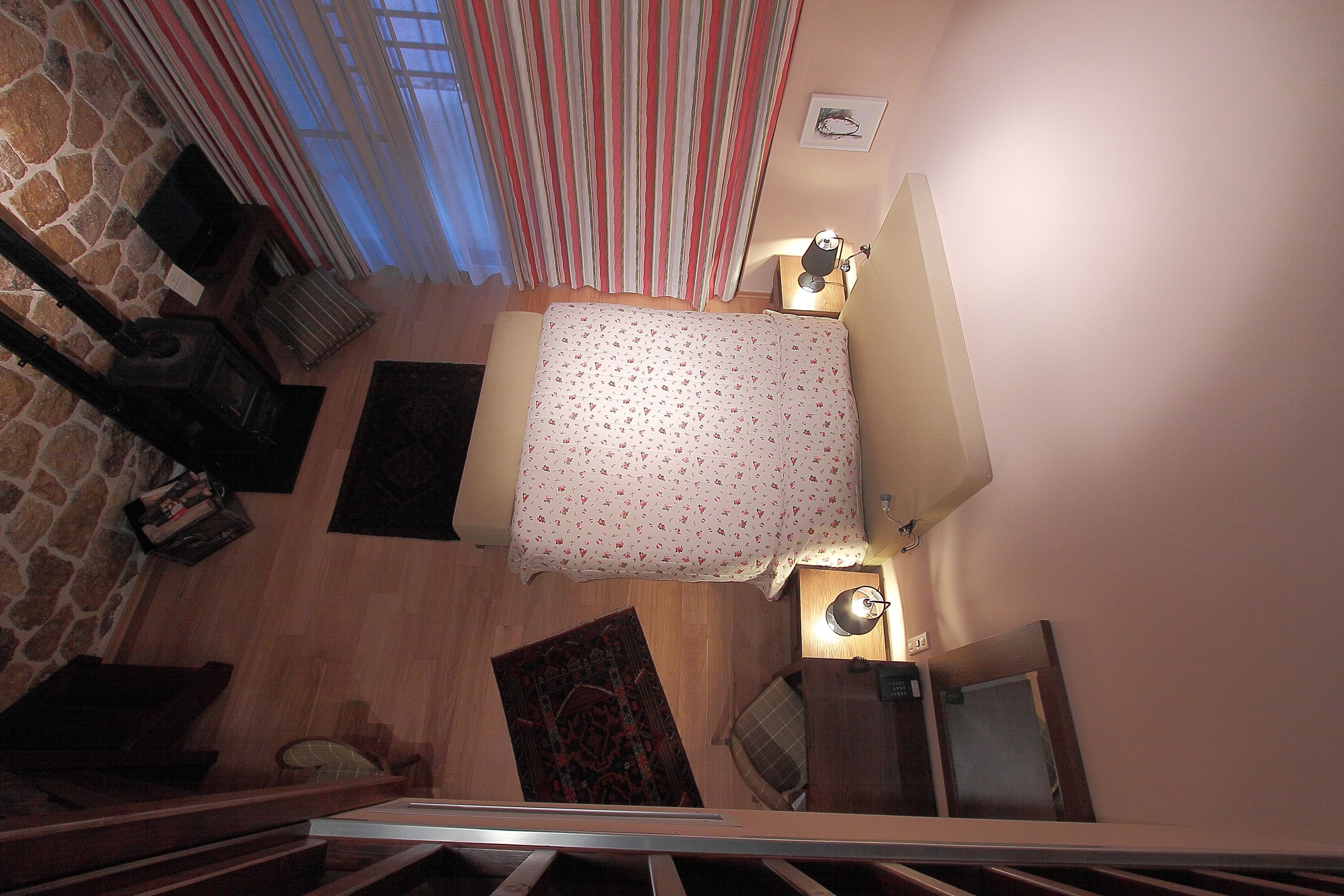Un dormitorio con cama, mesita de noche, lámpara, ventana con cortinas, pared de piedra y suelo de madera.