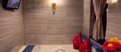 A hotel room with a bed, a wall-mounted TV, a wall-mounted light, a fan, a curtain, and a vase with flowers.