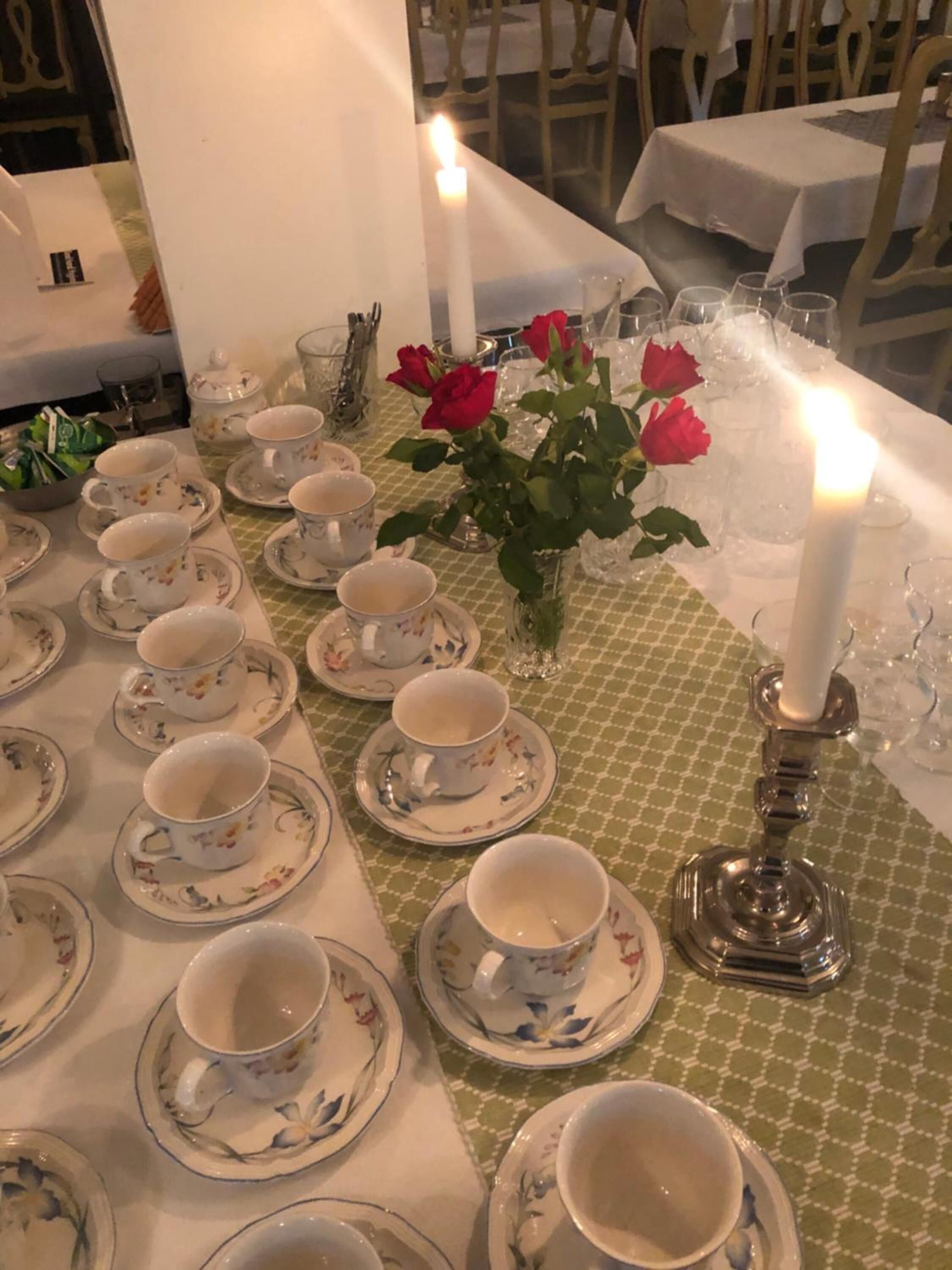 Une table à manger dressée avec des tasses et des soucoupes ornées de motifs floraux, un vase rempli de roses rouges et deux bougies allumées.