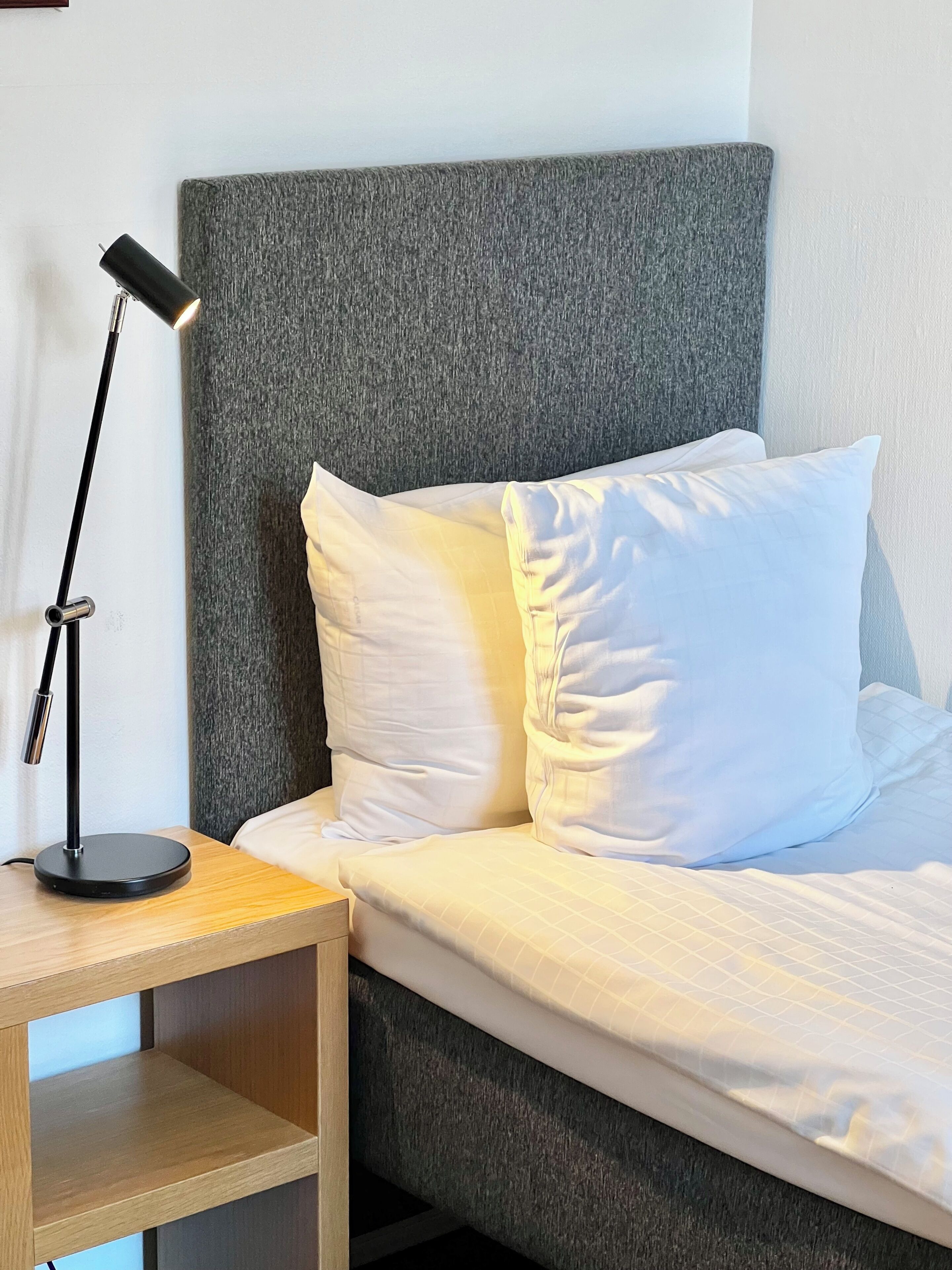 A single bed with white bedding, a grey headboard, and a black desk lamp on a wooden nightstand.