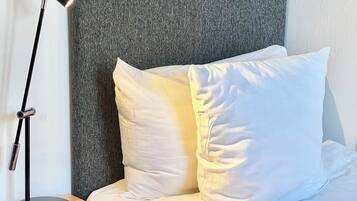 Uma cama de solteiro com roupa de cama branca, cabeceira cinzenta e abajur preto sobre uma mesa de cabeceira de madeira.