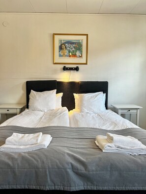 Un lit bien fait, avec des draps blancs, une couette grise et deux serviettes blanches posées sur le lit. Un tableau encadré est accroché au mur, au-dessus du lit.