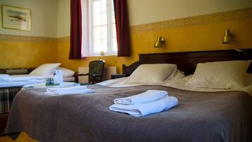 Habitación de hotel con una cama grande, una cama más pequeña, una silla, una mesa con lámpara y una ventana con cortinas.