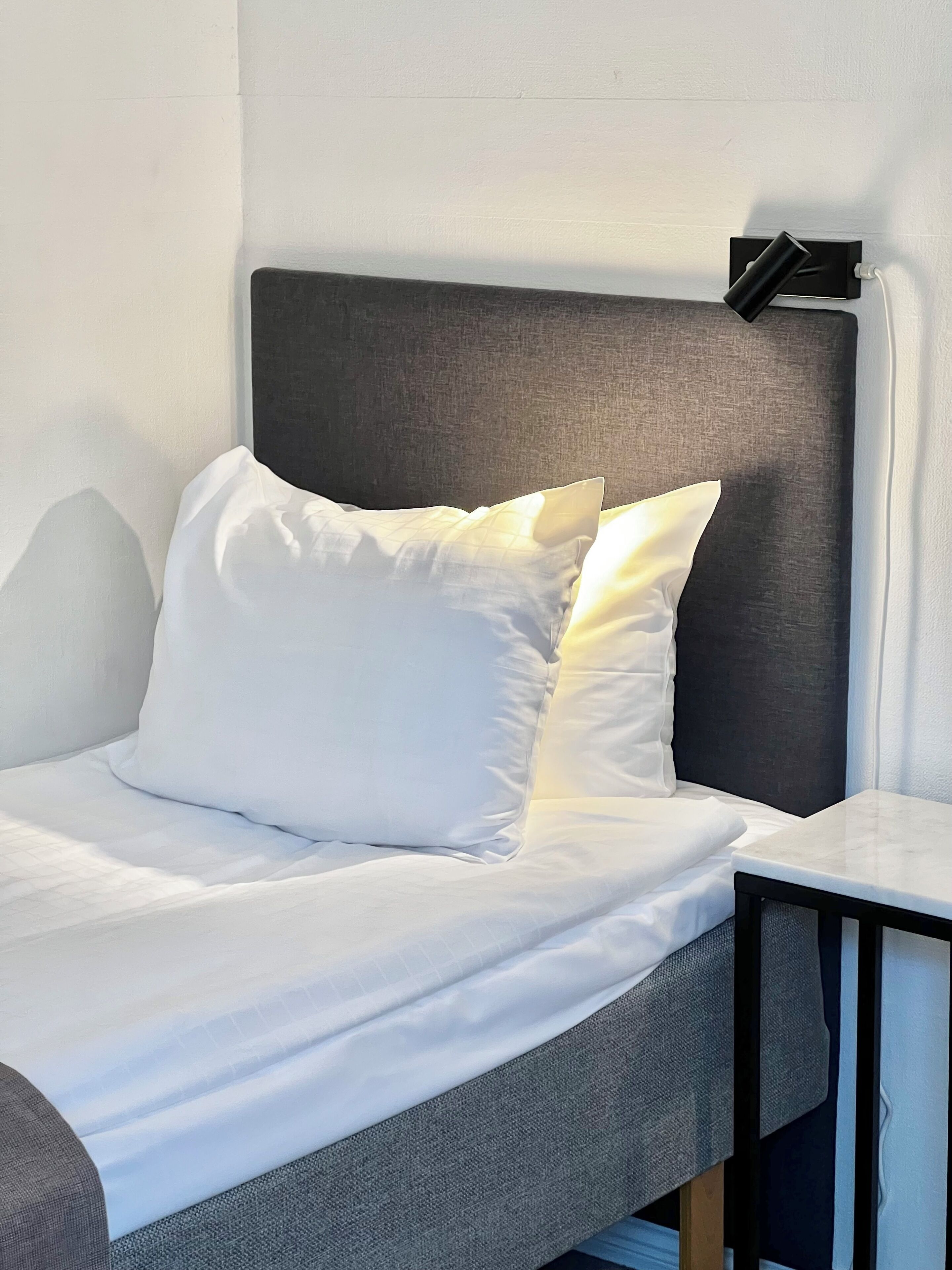 Una cama con ropa blanca, un cabecero gris y una mesita de noche con una lámpara.