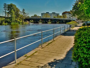 A riverside walkway with a metal railing, a stone bridge, and buildings in the background. - Värdshuset Hwitan (Falkenberg)
