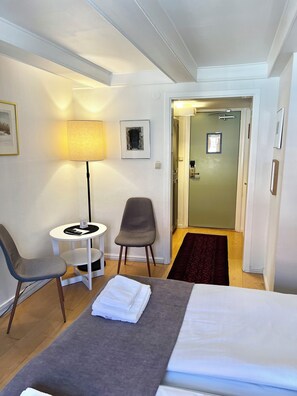 A hotel room with a bed, a desk with a lamp, two chairs, a small table, a framed picture on the wall, and a door leading to another room. - Värdshuset Hwitan (Falkenberg)