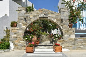 An entrance with a stone archway, a sign reading "Villa Katerina Rooms Studios," and potted plants with flowers. - Villa Katerina (Paros)
