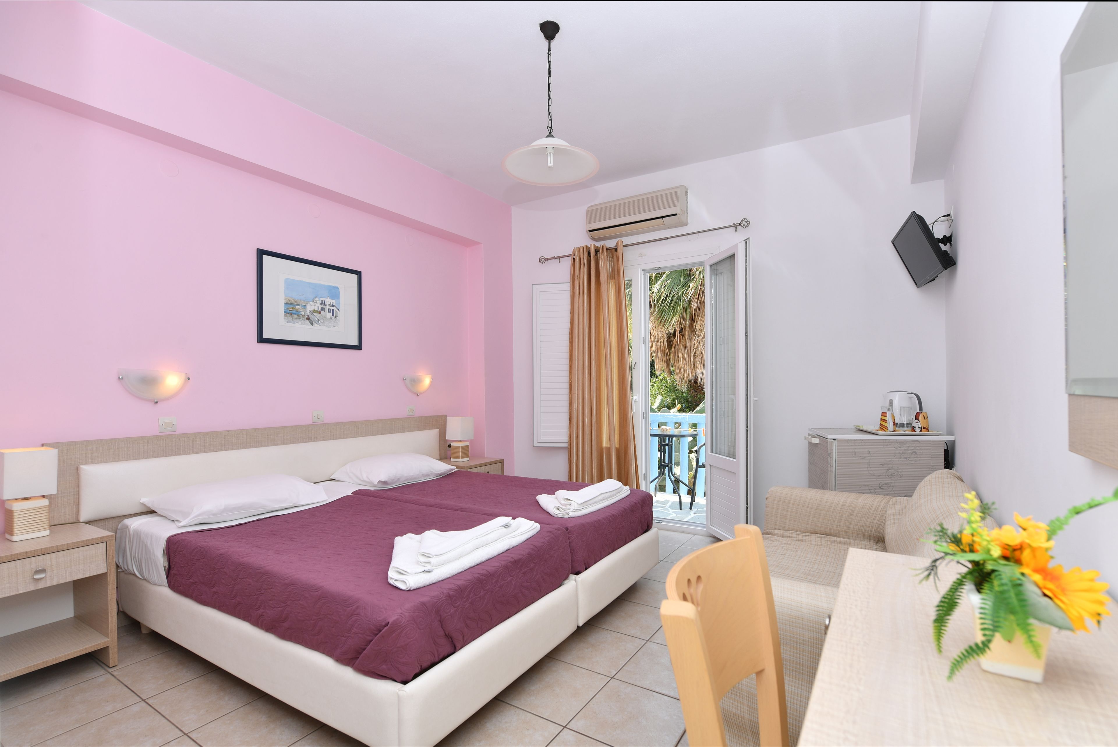 A bedroom with a bed, bedside tables, a desk, a chair, a table with a vase, and a view of the outside.