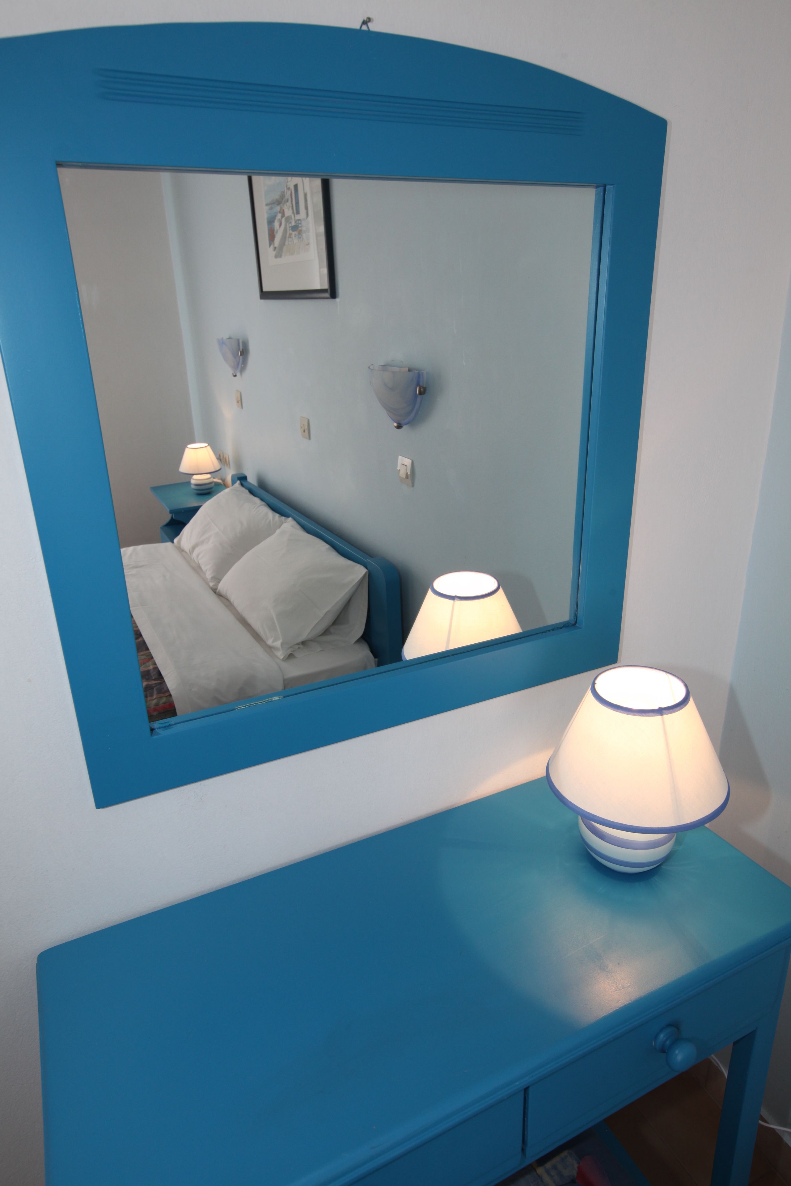 Een kamer met een bed, een nachtkastje met een lamp en een spiegel met een blauwe lijst.