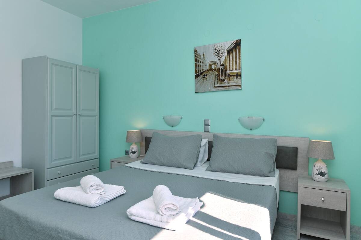 Una cama bien hecha con dos toallas enrolladas, un armario gris, una mesita de noche con una lámpara y un cuadro en la pared.