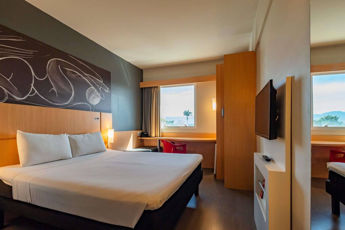 Quarto de hotel com uma cama grande, uma escrivaninha, uma televisão e uma janela com vista para a praia.