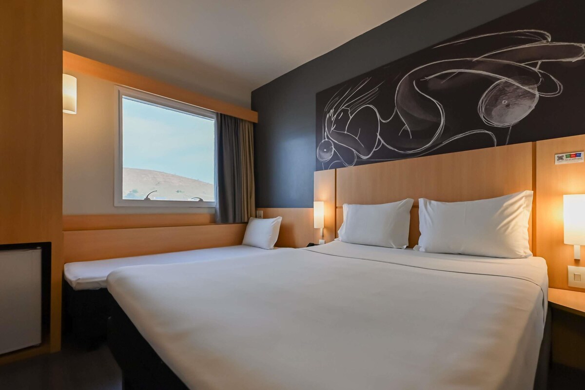 A hotel room with a large bed, a window with curtains, a wall-mounted artwork, and a nightstand with a lamp.
