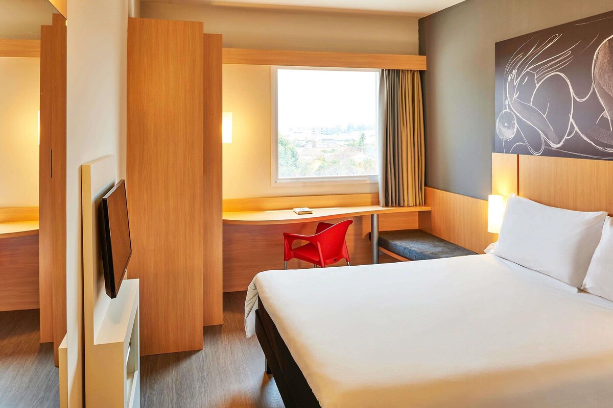 A hotel room with a bed, a desk, a red chair, and a TV.