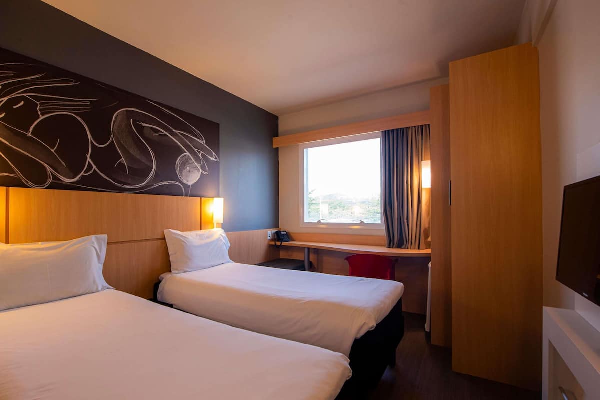 Quarto de hotel com duas camas, uma escrivaninha, televisão e janela com cortinas.
