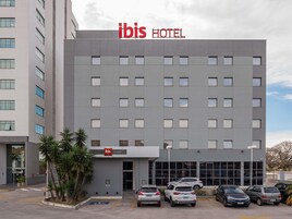 Un moderno edificio de hotel con un llamativo letrero 'ibis HOTEL', un estacionamiento con varios coches y un pequeño acceso con palmeras.