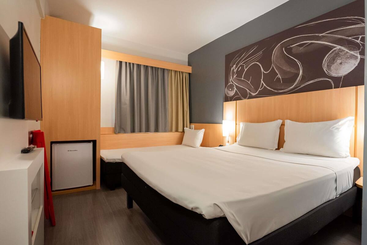Quarto de hotel com cama, televisão, pequena mesa de trabalho, janela com cortinas e uma obra de arte na parede.