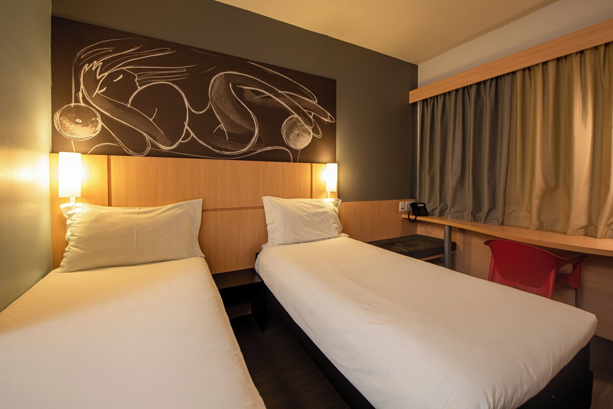 Een hotelkamer met twee enkele bedden, een houten hoofdbord met een wanddecoratie, een bureau met stoel en een raam met gordijnen.
