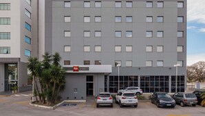 A modern hotel building with a prominent 'ibis HOTEL' sign, a parking lot with several cars, and a small entrance area with palm trees. - Ibis Guaratinguetá Aparecida (Guaratingueta)