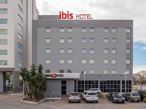 A modern hotel building with a prominent 'ibis HOTEL' sign, a parking lot with several cars, and a small entrance area with palm trees. - Ibis Guaratinguetá Aparecida (Guaratingueta)