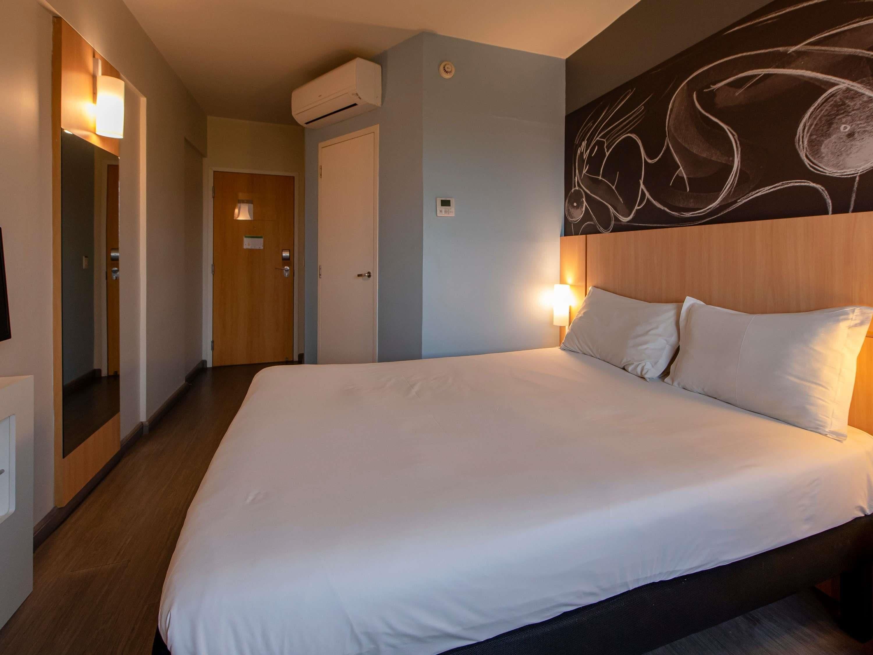 A hotel room with a large bed, a wooden headboard, a wall-mounted air conditioner, and a hallway with a door.