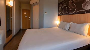 A hotel room with a large bed, a wooden headboard, a wall-mounted air conditioner, and a hallway with a door.