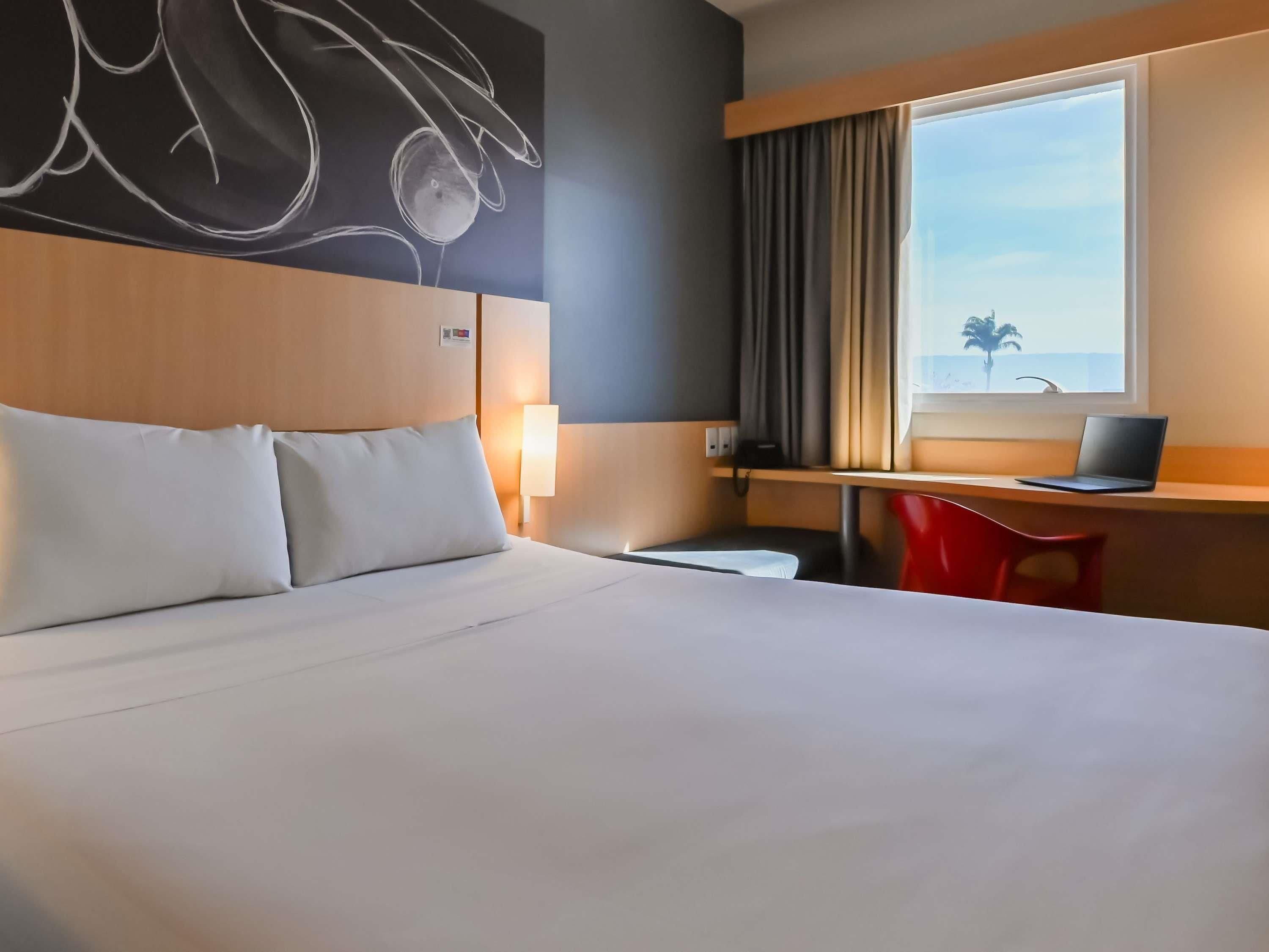 A hotel room with a bed, a desk with a laptop, a chair, a window with curtains, and a wall art piece.