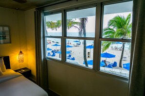 A hotel room with a large window overlooking a beach with blue umbrellas and palm trees. - Numero Uno Beach House (San Juan)