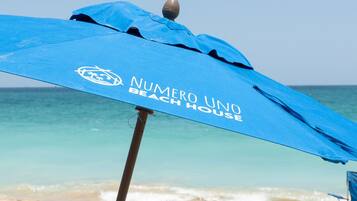 A blue beach umbrella with "NUMERO UNO BEACH HOUSE" written on it, set against a beach backdrop.