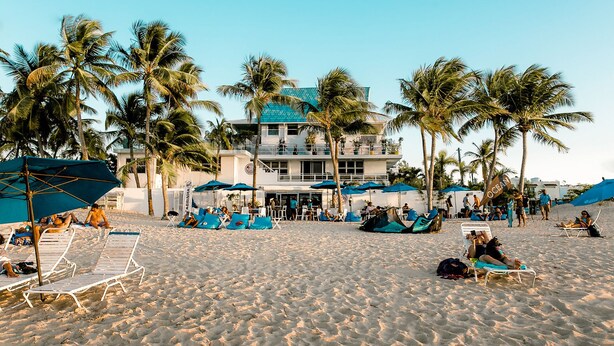 Un hotel frente a la playa con numerosas tumbonas y sombrillas, palmeras y un edificio de fachada azul.