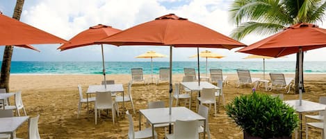 A beachside seating area with tables and chairs under umbrellas, overlooking the ocean.