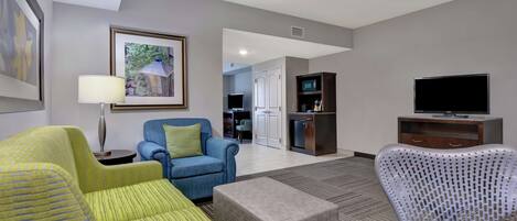 Habitación de hotel con sala de estar que incluye un sofá, un sillón, un reposapiés, televisión y una kitchenette.