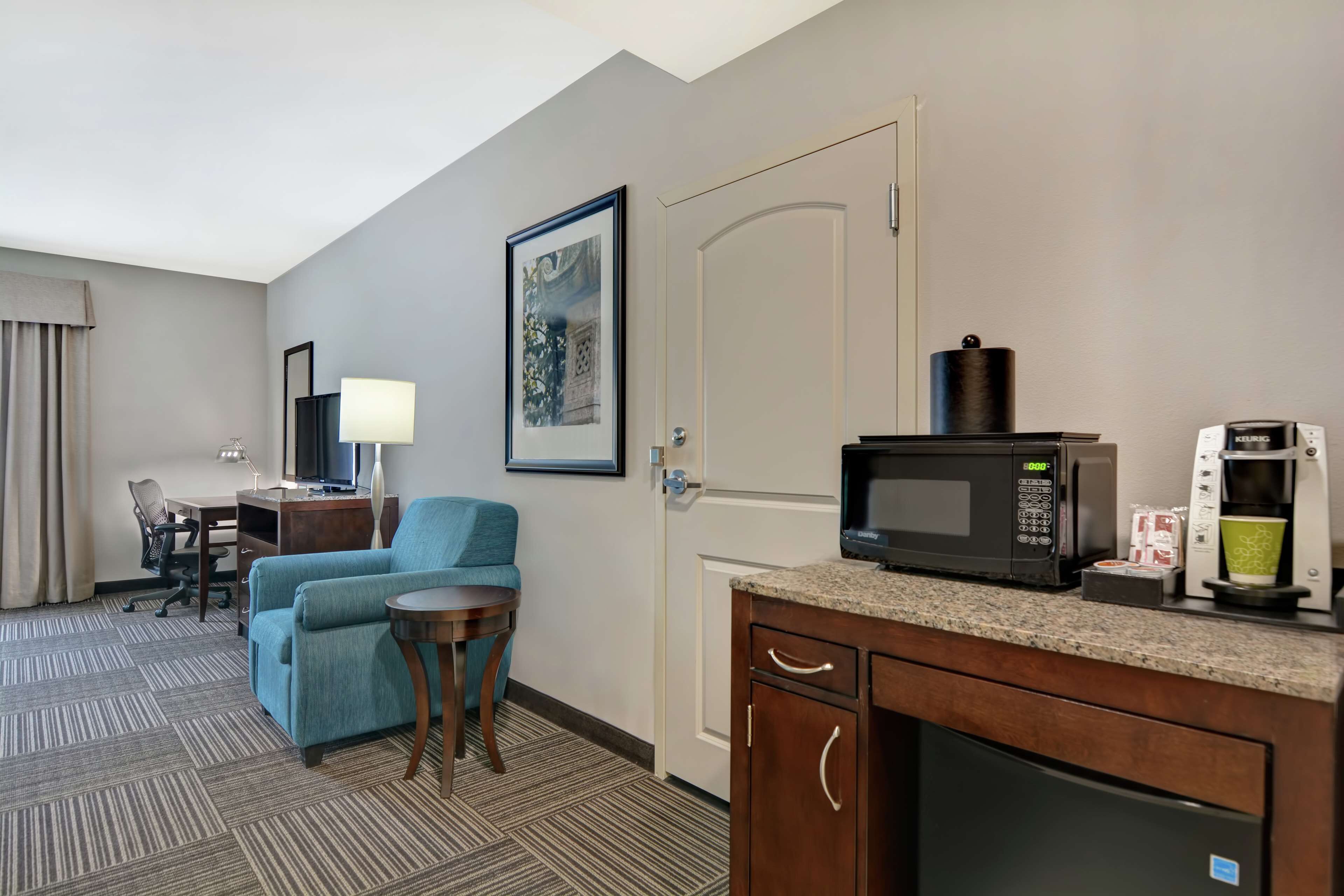 A hotel room with a kitchenette, a desk, a chair, a sofa, and a framed picture on the wall.