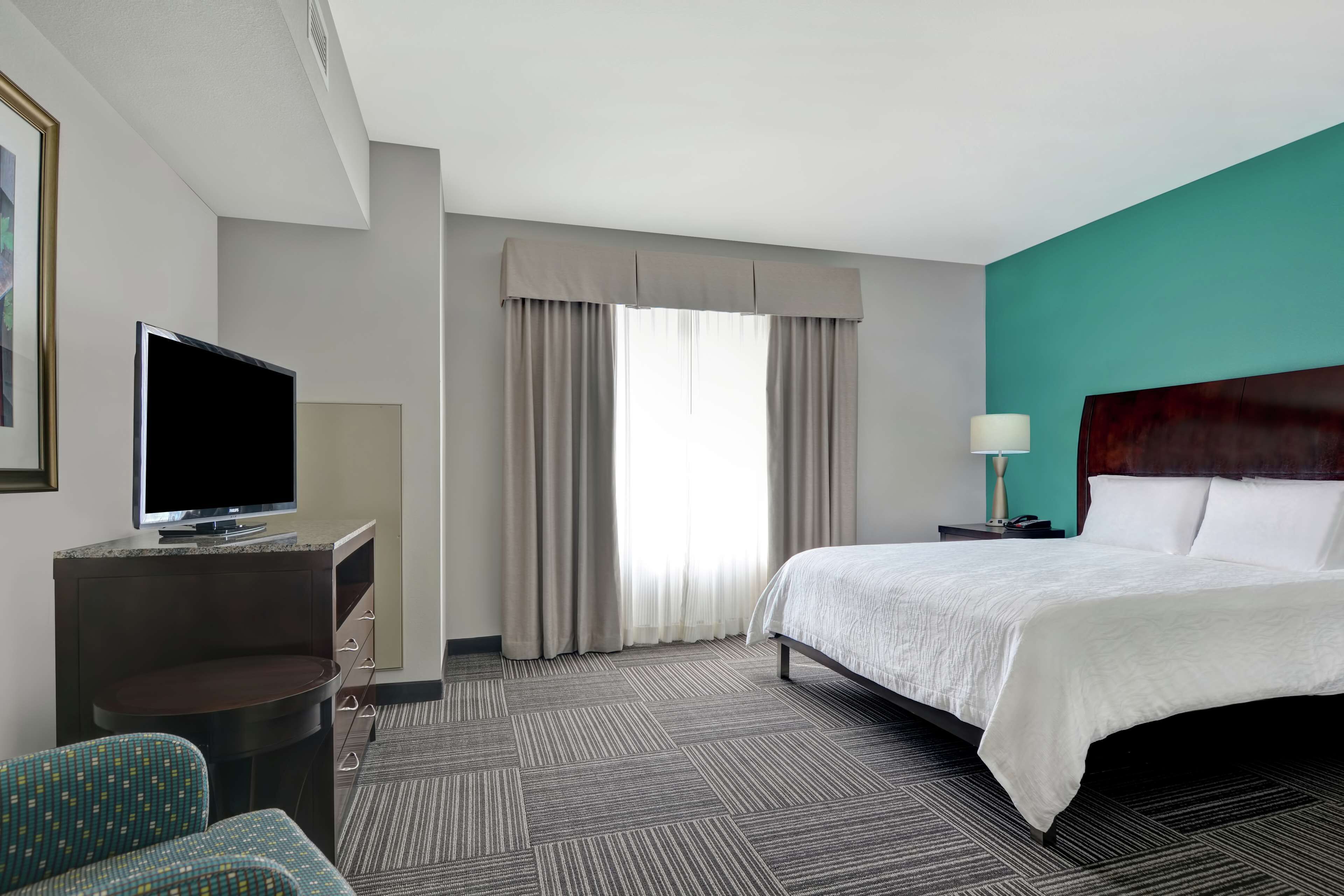 A hotel room with a bed, a desk with a TV, a chair, and a window with curtains.