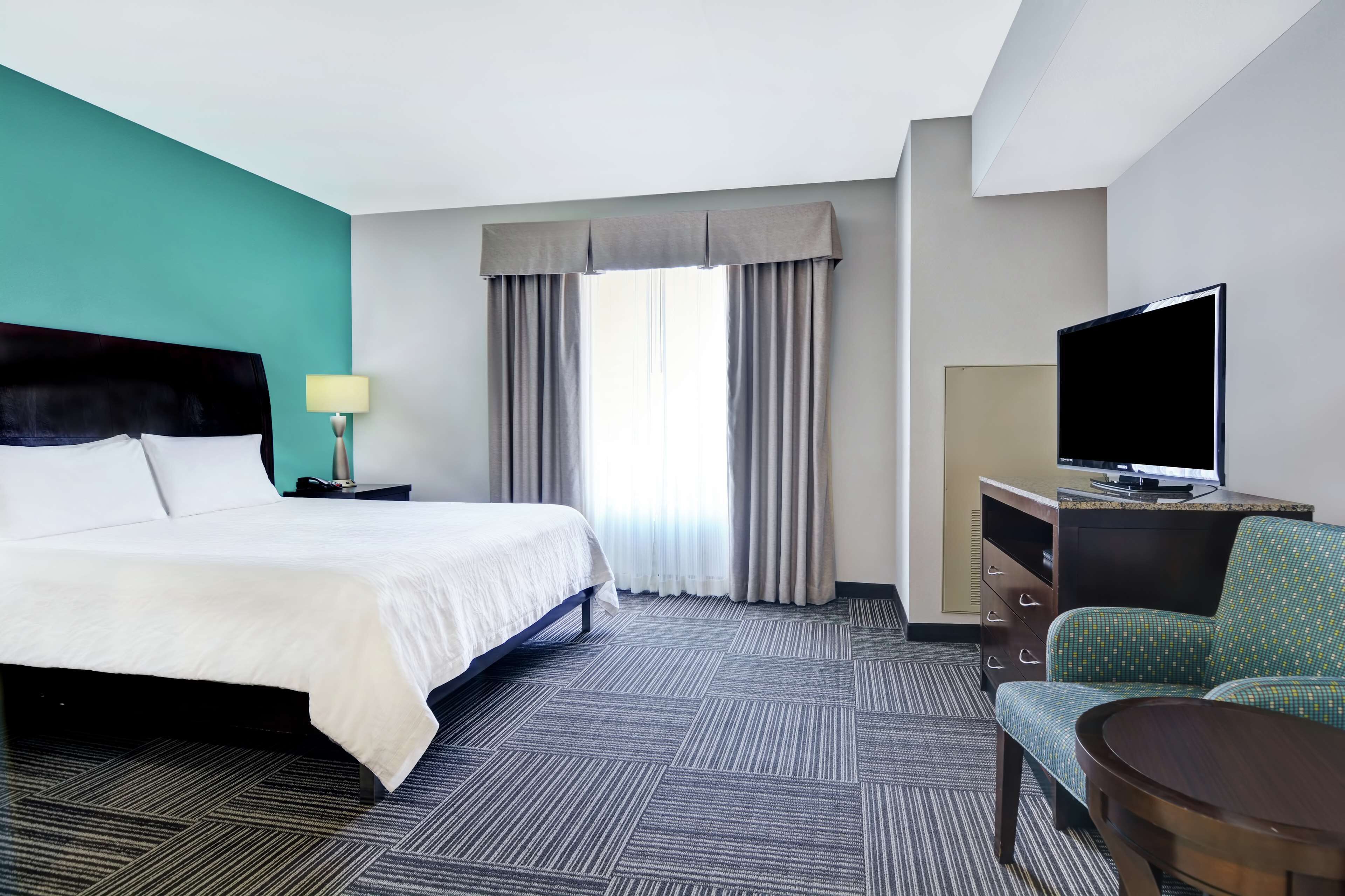 A hotel room with a bed, a desk with a TV, a chair, and a window with curtains.