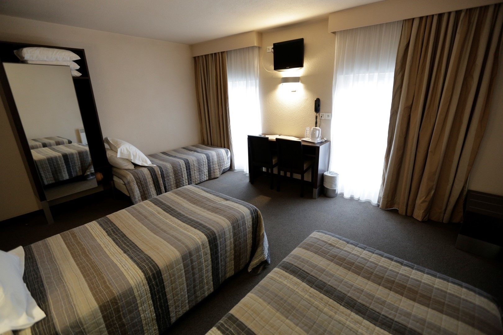 A hotel room with two single beds, a desk with a chair, a TV mounted on the wall, and a window with curtains.