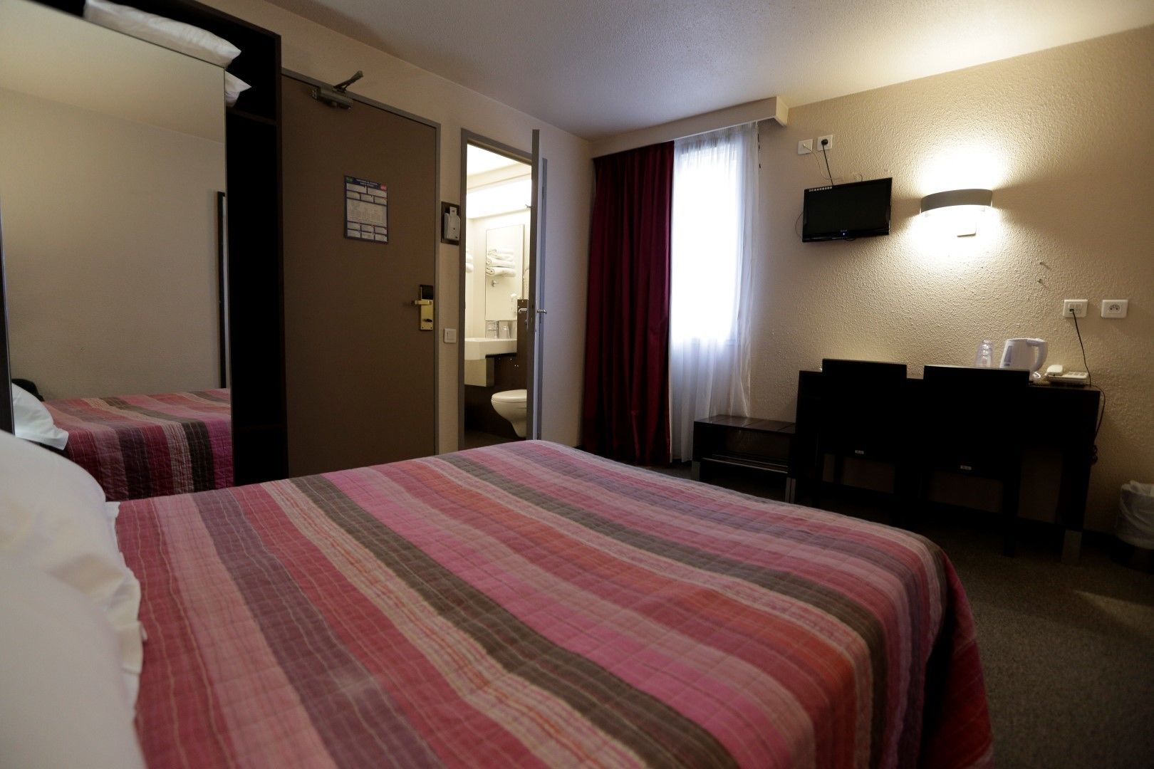 A hotel room with two beds, a desk, a TV, and a bathroom visible through an open door.