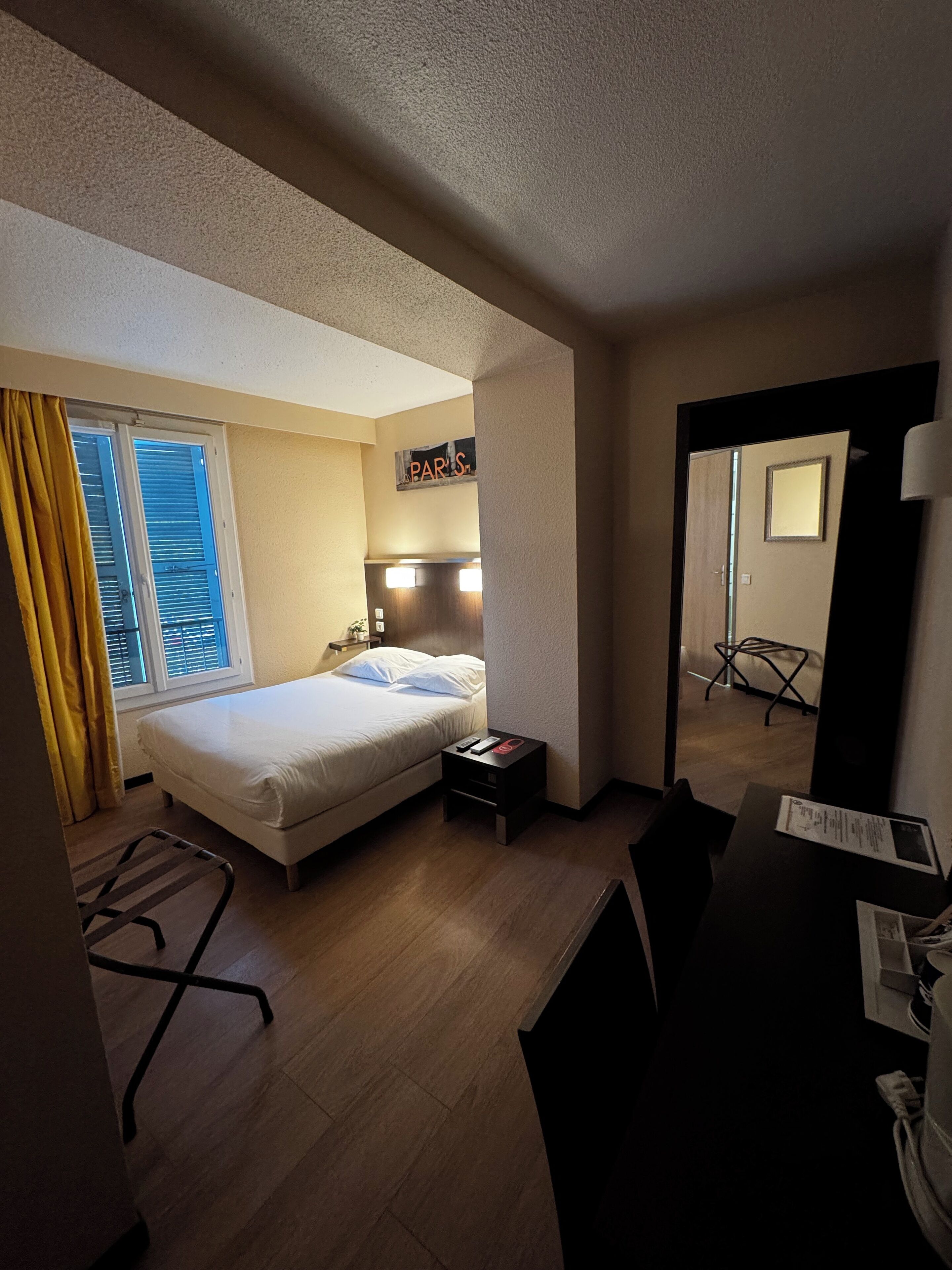 Habitación de hotel con cama, mesita de noche, escritorio, silla, espejo y ventana con cortinas.