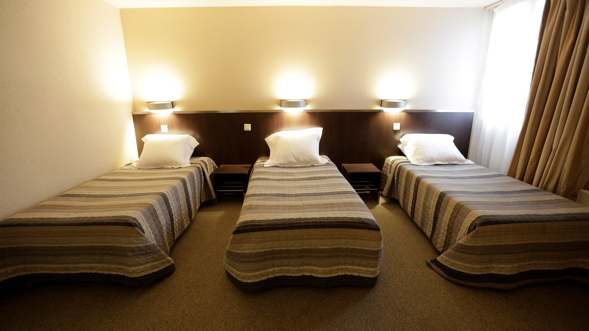 A hotel room with three single beds, each with a striped bedspread and white pillows. There are wall-mounted lights above each bed.