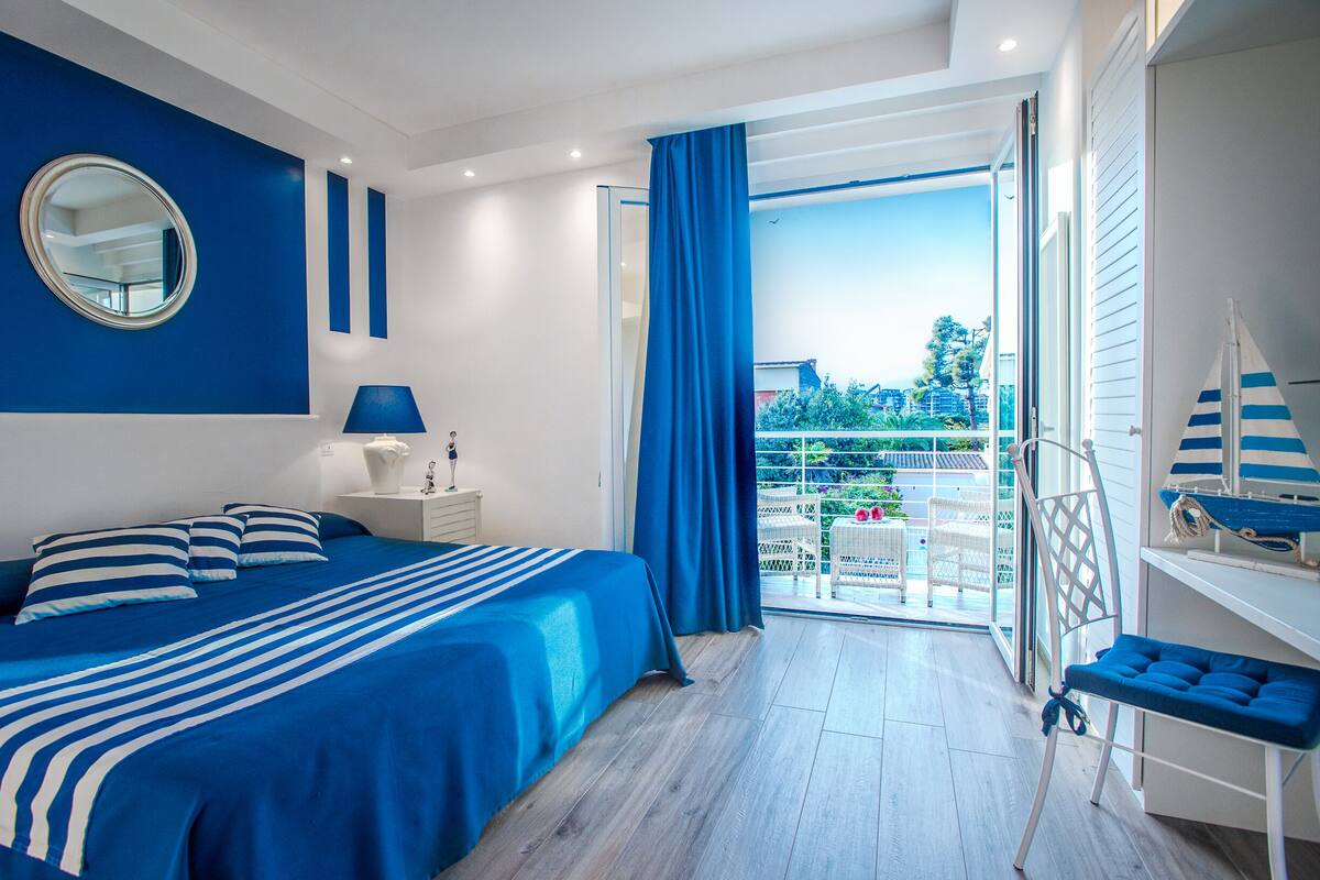 A bedroom with a large bed, a nightstand, a mirror, and a view of the outdoors.