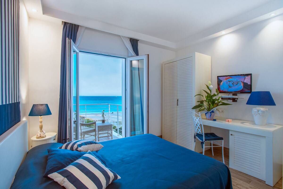 A bedroom with a bed, a desk, a chair, a TV, and a view of the ocean.