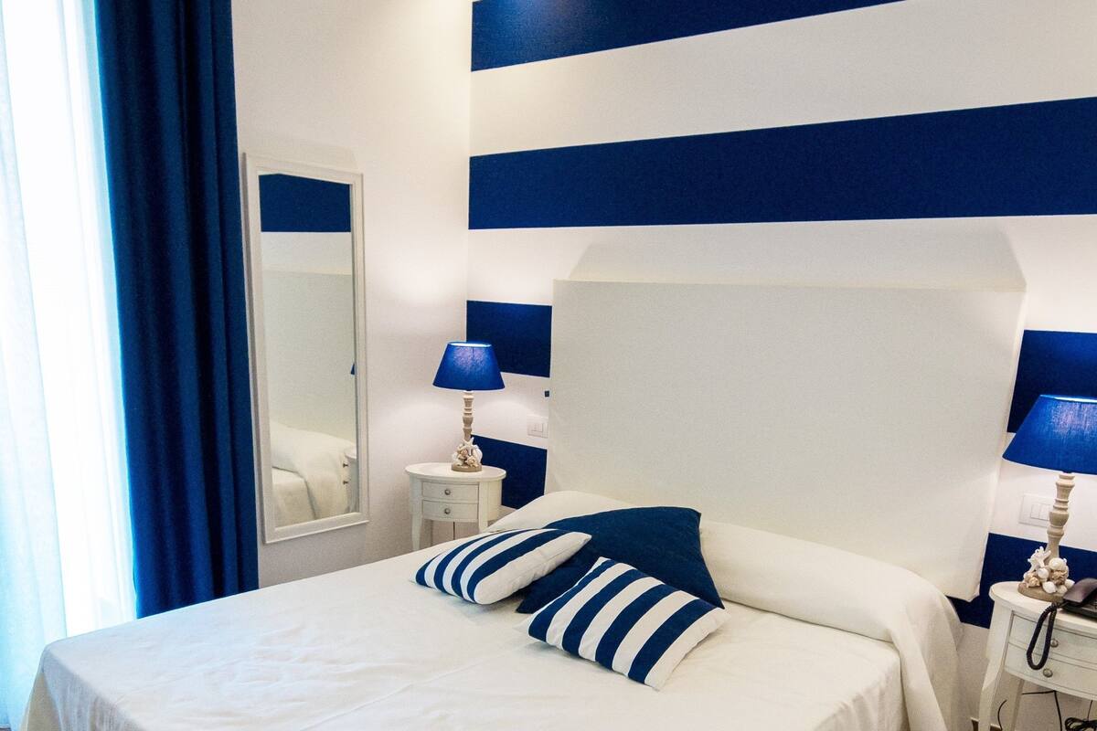 A neatly arranged bedroom with a large bed, two bedside tables, a mirror, and a blue-striped wall.