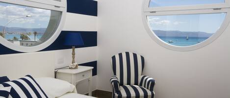 A room with a bed, a chair, a nightstand, and two windows offering a view of the sea and mountains.